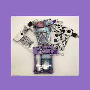 Könnte beinhalten: Vier transparente Reißverschlusstaschen mit verschiedenen Nightmare Before Christmas-Charakterdrucken. Die Taschen haben einen violetten Besatz und einen violetten Knopfverschluss. Eine Tasche enthält eine kleine Flasche Handdesinfektionsmittel und einen Lippenbalsam.