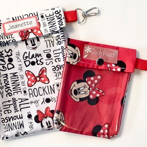 Könnte beinhalten: Zwei rote und weiße Minnie Mouse-thematische Reißverschlusstaschen mit durchsichtigen Vinyl-Fronten. Die größere Tasche hat einen weißen Hintergrund mit schwarzem Text, der "Glam Dots" und "Rockin' my dots" sagt. Die kleinere Tasche hat einen roten Hintergrund mit weißen Punkten und ein klares Etikett, auf dem "Ouch Pouch" steht.