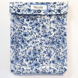 May include: A blue floral print case with a flap closure secured by three blue buttons. The case features a label that reads "Nicole" in a white font. The floral pattern is a mix of blue hues on a white background.