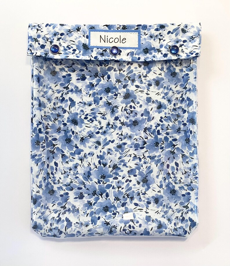 May include: A blue floral print case with a flap closure secured by three blue buttons. The case features a label that reads "Nicole" in a white font. The floral pattern is a mix of blue hues on a white background.