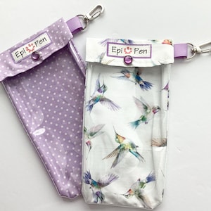 May include: Two EpiPen cases. One is lavender with white polka dots, the other is white with a hummingbird pattern. Both have a clear plastic front, a snap closure, and a silver carabiner clip. The text "Epi Pen" is printed on the top.