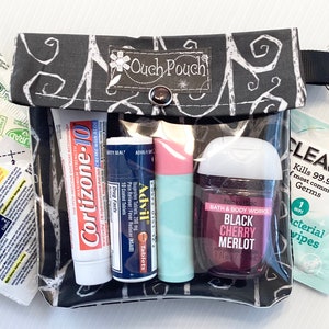 May include: A black and white patterned pouch with a clear plastic window. The pouch contains various items, including a tube of Cortizone-10 cream, a tube of Advil tablets, a small bottle of hand sanitizer, a tube of lip balm, and a packet of antibacterial wipes. The pouch has a zipper closure and a black strap with a silver hook.