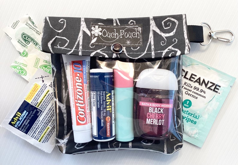 May include: A black and white patterned pouch with a clear plastic window. The pouch contains various items, including a tube of Cortizone-10 cream, a tube of Advil tablets, a small bottle of hand sanitizer, a tube of lip balm, and a packet of antibacterial wipes. The pouch has a zipper closure and a black strap with a silver hook.
