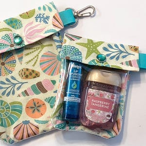 May include: Two clear pouches with a seashell pattern, teal accents, and silver carabiner clips. One pouch contains a blue lip balm and a raspberry tangerine hand sanitizer. The other pouch is empty.
