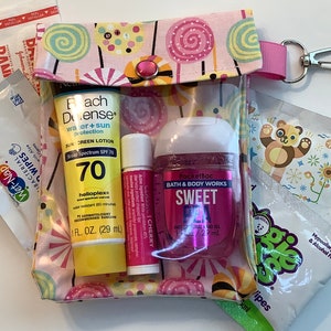 Może przedstawiać: Przezroczysta kosmetyczka zapinana na zamek z r&oacute;żowo-białym nadrukiem cukierk&oacute;w, zawierająca krem ​​przeciwsłoneczny, balsam do ust, środek do dezynfekcji rąk i chusteczki. Kosmetyczka ma r&oacute;żowy pasek i srebrny klips. Na butelce z kremem przeciwsłonecznym widnieje napis "SPF 70".