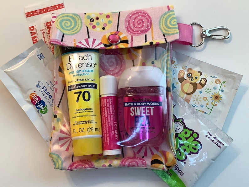 Może przedstawiać: Przezroczysta kosmetyczka zapinana na zamek z r&oacute;żowo-białym nadrukiem cukierk&oacute;w, zawierająca krem ​​przeciwsłoneczny, balsam do ust, środek do dezynfekcji rąk i chusteczki. Kosmetyczka ma r&oacute;żowy pasek i srebrny klips. Na butelce z kremem przeciwsłonecznym widnieje napis "SPF 70".