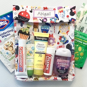 Könnte beinhalten: Eine weiße Stofftasche mit einem bunten Druck von Cupcakes, Donuts und Mickey-Maus-Ohren. Die Tasche ist mit "Abigail" beschriftet und enthält verschiedene Artikel, darunter Sonnencreme, Händedesinfektionsmittel, Feuchttücher und Schmerzmittel.