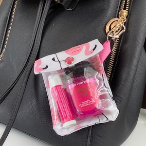 May include: A clear plastic zippered pouch with a flamingo print and a pink button closure. The pouch is attached to a black backpack with a gold zipper. Inside the pouch are a pink hand sanitizer bottle with the text "Sweet Pea PocketBac Anti-Bacterial Hand Gel with Essential Oils" and a pink lip balm.