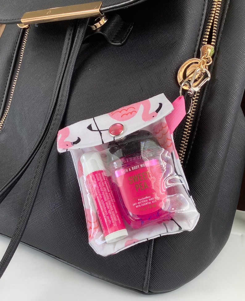 May include: A clear plastic zippered pouch with a flamingo print and a pink button closure. The pouch is attached to a black backpack with a gold zipper. Inside the pouch are a pink hand sanitizer bottle with the text "Sweet Pea PocketBac Anti-Bacterial Hand Gel with Essential Oils" and a pink lip balm.