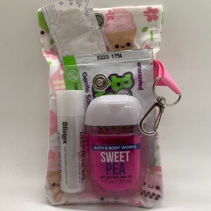 Może przedstawiać: Mała, materiałowa torebka z r&oacute;żowym paskiem i srebrnym zapięciem, wypełniona artykułami do pielęgnacji osobistej. Torebka ma białe tło z r&oacute;żowymi kwiatami i rożkami lod&oacute;w. Zawartość: balsam do ust, środek do dezynfekcji rąk i saszetka. Środek do dezynfekcji rąk jest oznaczony jako "Sweet Pea".