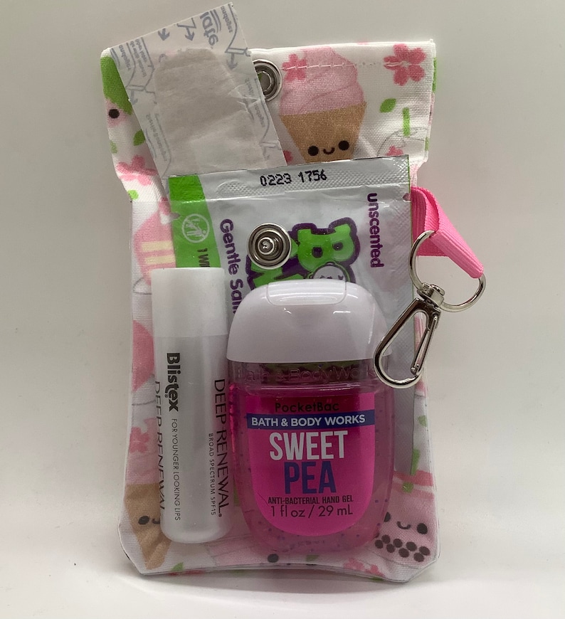 Może przedstawiać: Mała, materiałowa torebka z r&oacute;żowym paskiem i srebrnym zapięciem, wypełniona artykułami do pielęgnacji osobistej. Torebka ma białe tło z r&oacute;żowymi kwiatami i rożkami lod&oacute;w. Zawartość: balsam do ust, środek do dezynfekcji rąk i saszetka. Środek do dezynfekcji rąk jest oznaczony jako "Sweet Pea".