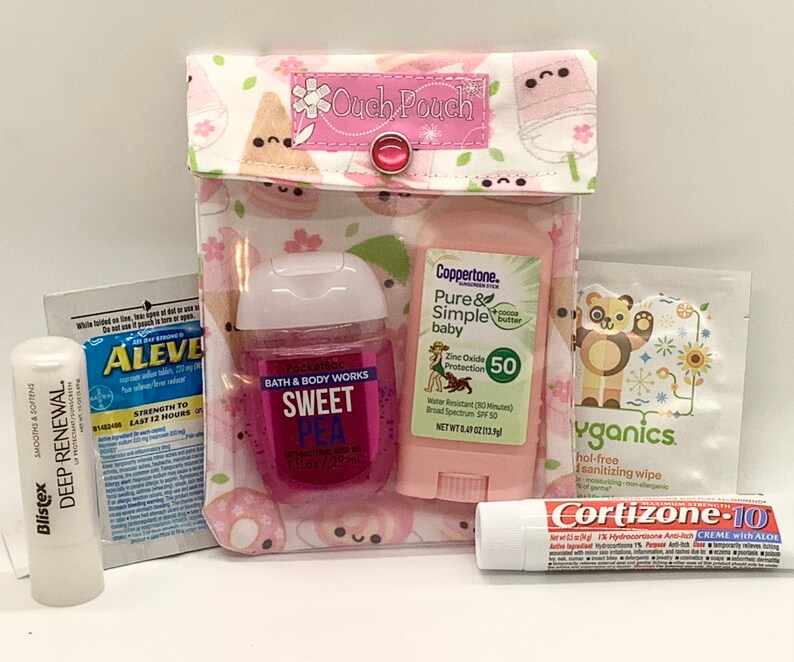 Może przedstawiać: Przezroczysty, r&oacute;żowy "Ouch Pouch" z zapięciem na zatrzask, zawierający r&oacute;żne produkty zdrowotne i kosmetyczne. Zawiera balsam do ust Blistex, środek przeciwb&oacute;lowy Aleve, środek do dezynfekcji rąk, krem ​​przeciwsłoneczny, chusteczkę dezynfekującą i krem ​​Cortizone-10.