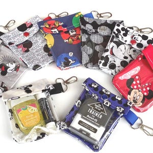 May include: A collection of small, rectangular pouches featuring various Disney character prints, including Mickey and Minnie Mouse. Each pouch has a snap closure, a metal lobster clasp, and a clear plastic front window. Some pouches contain items like hand sanitizer, lip balm, or an Auvi-Q trainer.
