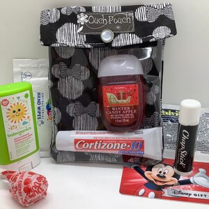 Może przedstawiać: Czarna i biała torebka z napisem "Ouch Pouch" zawierająca r&oacute;żne przedmioty. Zawiera środek do dezynfekcji rąk, sztyft do opalania, balsam do ust, chusteczki nawilżane, kartę podarunkową Disneya i lizaka.