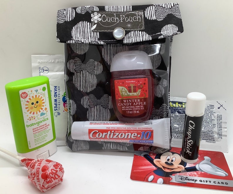 Może przedstawiać: Czarna i biała torebka z napisem "Ouch Pouch" zawierająca r&oacute;żne przedmioty. Zawiera środek do dezynfekcji rąk, sztyft do opalania, balsam do ust, chusteczki nawilżane, kartę podarunkową Disneya i lizaka.