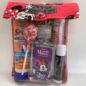 May include: A red and white Minnie Mouse themed pouch with a clear plastic window. The pouch contains a variety of items, including sunscreen, a lollipop, hand sanitizer, lip balm, and a tube of cream. The text on the pouch reads "Ouch Pouch".