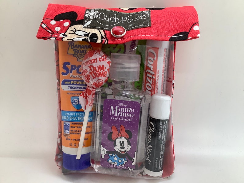 May include: A red and white Minnie Mouse themed pouch with a clear plastic window. The pouch contains a variety of items, including sunscreen, a lollipop, hand sanitizer, lip balm, and a tube of cream. The text on the pouch reads "Ouch Pouch".
