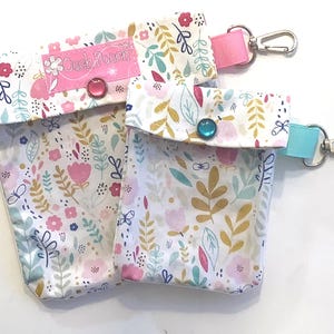 May include: Two small floral print pouches with snap closures and attached key rings. The pouches are white with pink, blue, and yellow floral designs. The larger pouch has a pink label that says "Ouch Pouch".