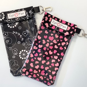 May include: Two black fabric cases with clear plastic windows and zipper closures. The cases have a black and white floral pattern and a pink and black heart pattern. Both cases have a black label with white text that reads "Epi Pen".