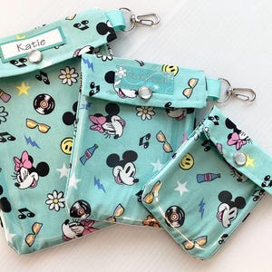 Może przedstawiać: Trzy turkusowe saszetki zapinane na zamek błyskawiczny z postaciami Myszki Miki i Minnie. Największa saszetka ma napisane na niej imię "Katie". Wszystkie trzy saszetki mają srebrny brelok.