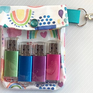 May include: A white fabric pouch with a rainbow and heart pattern, featuring a turquoise button and a teal strap with a silver key ring. The pouch contains four USB flash drives: green, blue, pink, and purple.