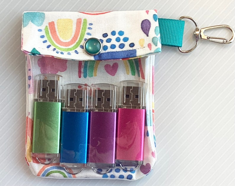 May include: A white fabric pouch with a rainbow and heart pattern, featuring a turquoise button and a teal strap with a silver key ring. The pouch contains four USB flash drives: green, blue, pink, and purple.