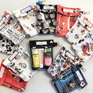 May include: A collection of small, rectangular, clear vinyl pouches featuring various Disney-themed patterns, including Mickey and Minnie Mouse. Each pouch has a flap with a personalized name label, a snap closure, and a metal swivel clip attachment.