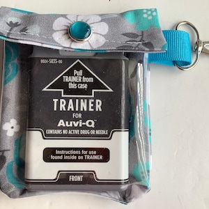 May include: A gray and teal floral fabric case with a clear plastic window. The case contains a black Auvi-Q trainer with the text "Pull TRAINER from this case", "TRAINER FOR Auvi-Q", "CONTAINS NO ACTIVE DRUG OR NEEDLE", "Instructions for use found inside on TRAINER", and "FRONT". The case has a blue strap with a silver clip.