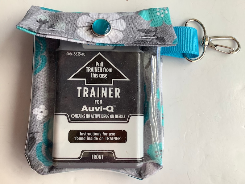 May include: A gray and teal floral fabric case with a clear plastic window. The case contains a black Auvi-Q trainer with the text "Pull TRAINER from this case", "TRAINER FOR Auvi-Q", "CONTAINS NO ACTIVE DRUG OR NEEDLE", "Instructions for use found inside on TRAINER", and "FRONT". The case has a blue strap with a silver clip.