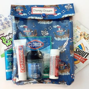 May include: A clear, zippered pouch with a blue nautical-themed fabric featuring cartoon boats and anchors. The pouch contains various travel-sized toiletries, including lip balm, hand sanitizer, and a small bottle of Cortizone-10. The text "Disney Dream" is on the pouch.