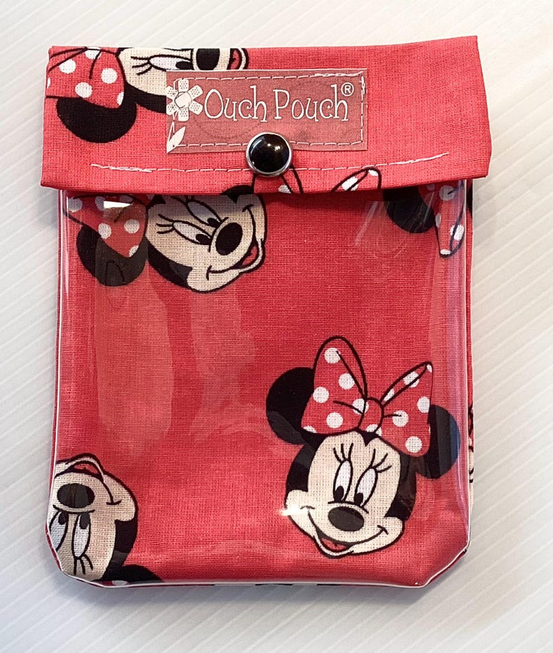 May include: A red fabric pouch with a clear plastic front. The pouch has a white button closure and features a repeating pattern of Minnie Mouse. The pouch has a label that reads "Ouch Pouch".