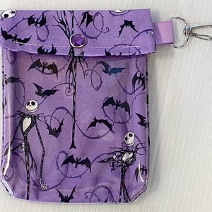 Puede incluir: Una bolsa de cremallera morada con un panel frontal transparente que presenta un patr&oacute;n repetido de murci&eacute;lagos negros y un personaje de Pesadilla Antes de Navidad. La bolsa tiene un cierre de presi&oacute;n negro y un llavero de metal negro y plateado.