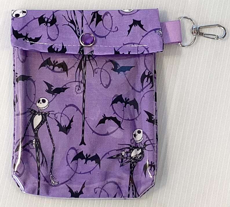 Puede incluir: Una bolsa de cremallera morada con un panel frontal transparente que presenta un patr&oacute;n repetido de murci&eacute;lagos negros y un personaje de Pesadilla Antes de Navidad. La bolsa tiene un cierre de presi&oacute;n negro y un llavero de metal negro y plateado.