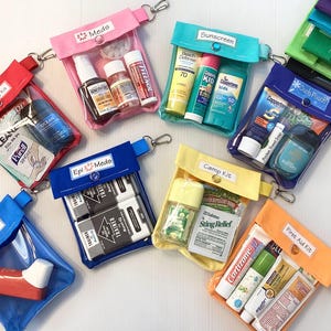 May include: Nine clear plastic zippered pouches with different colored fabric exteriors and labels. The pouches contain various first aid and travel items, including sunscreen, hand sanitizer, pain relief, and an inhaler. The labels on the pouches read: "Emergency Kit", "Mede", "Epi 3 Meds", "Camp Kit", "First Aid Kit", "Ouch Pouch", "Sunscreen", and "Inhaler".