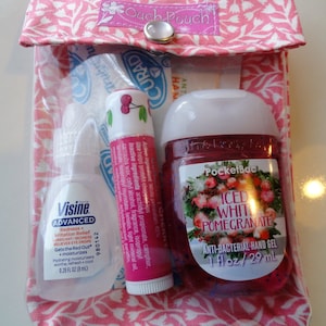 K&ouml;nnte beinhalten: Eine rosa Stofftasche mit einem durchsichtigen Plastikfenster, in dem sich drei kleine Flaschen befinden. Die Flaschen enthalten Augentropfen, Lippenbalsam und H&auml;ndedesinfektionsmittel. Der Lippenbalsam ist Kirschgeschmack und das H&auml;ndedesinfektionsmittel ist PocketBac Iced White Pomegranate. Die Tasche hat einen rosa Knopfverschluss und der Text "Ouch Pouch" ist auf der Vorderseite aufgedruckt.