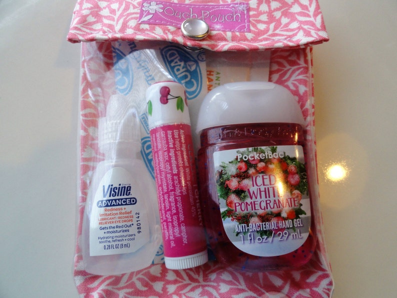 K&ouml;nnte beinhalten: Eine rosa Stofftasche mit einem durchsichtigen Plastikfenster, in dem sich drei kleine Flaschen befinden. Die Flaschen enthalten Augentropfen, Lippenbalsam und H&auml;ndedesinfektionsmittel. Der Lippenbalsam ist Kirschgeschmack und das H&auml;ndedesinfektionsmittel ist PocketBac Iced White Pomegranate. Die Tasche hat einen rosa Knopfverschluss und der Text "Ouch Pouch" ist auf der Vorderseite aufgedruckt.