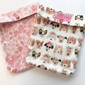 May include: Two rectangular pouches with snap closures. One pouch features a pink floral pattern. The other pouch has a white background with a repeating print of dog faces wearing flower crowns and a pink label that reads Ouch Pouch.