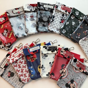 K&ouml;nnte beinhalten: Eine Sammlung von 12 bunten Mickey-Mouse-Rei&szlig;verschlusstaschen mit durchsichtigen Plastikfenstern. Die Taschen haben verschiedene Mickey-Mouse- und Minnie-Mouse-Muster und sind mit Namen wie Hayden, Joshua, Rebecca und Disney Dream personalisiert. Jede Tasche hat einen schwarzen Knopf und einen silbernen Schl&uuml;sselring.
