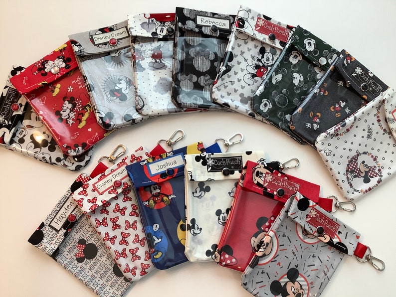 K&ouml;nnte beinhalten: Eine Sammlung von 12 bunten Mickey-Mouse-Rei&szlig;verschlusstaschen mit durchsichtigen Plastikfenstern. Die Taschen haben verschiedene Mickey-Mouse- und Minnie-Mouse-Muster und sind mit Namen wie Hayden, Joshua, Rebecca und Disney Dream personalisiert. Jede Tasche hat einen schwarzen Knopf und einen silbernen Schl&uuml;sselring.