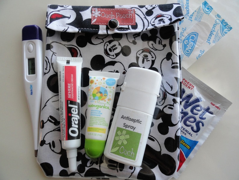 May include: A black and white Mickey Mouse print pouch containing a thermometer, Orajel severe toothache cream, babyganics soothing protective ointment, antiseptic spray, and Wet Ones antibacterial wipes. The pouch is labeled "Ouch Pouch".