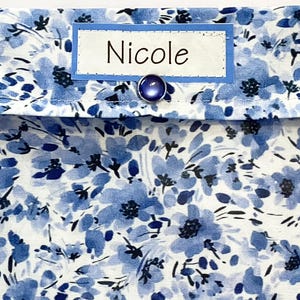 May include: A blue and white floral pattern fabric with the name "Nicole" on a label. The fabric is adorned with three dark blue, round buttons. The design features a delicate floral print in shades of blue on a white background.