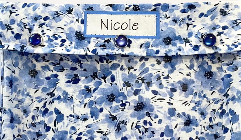 May include: A blue and white floral pattern fabric with the name "Nicole" on a label. The fabric is adorned with three dark blue, round buttons. The design features a delicate floral print in shades of blue on a white background.