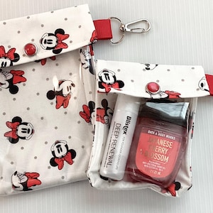 May include: Two clear plastic zippered pouches with white fabric exteriors featuring a repeating pattern of red and white polka dots and cartoon Minnie Mouse heads. The pouches have red fabric straps with silver key rings. The smaller pouch contains a clear bottle of hand sanitizer with the text "Bath & Body Works Japanese Cherry Blossom" and a clear tube of hand cream with the text "Dlistex Deep Renewal".
