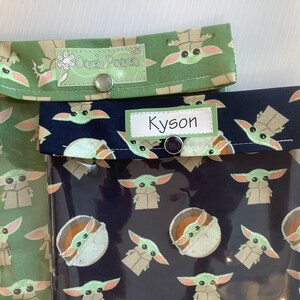 Puede incluir: Dos bolsas de aperitivos reutilizables con un frente de vinilo transparente y una parte posterior de tela. Las bolsas est&aacute;n decoradas con un patr&oacute;n repetido de un personaje alien&iacute;gena verde. Una bolsa tiene una parte posterior de tela verde con una etiqueta que dice "Ouch Pouch" y la otra bolsa tiene una parte posterior de tela negra con una etiqueta que dice "Kyson".