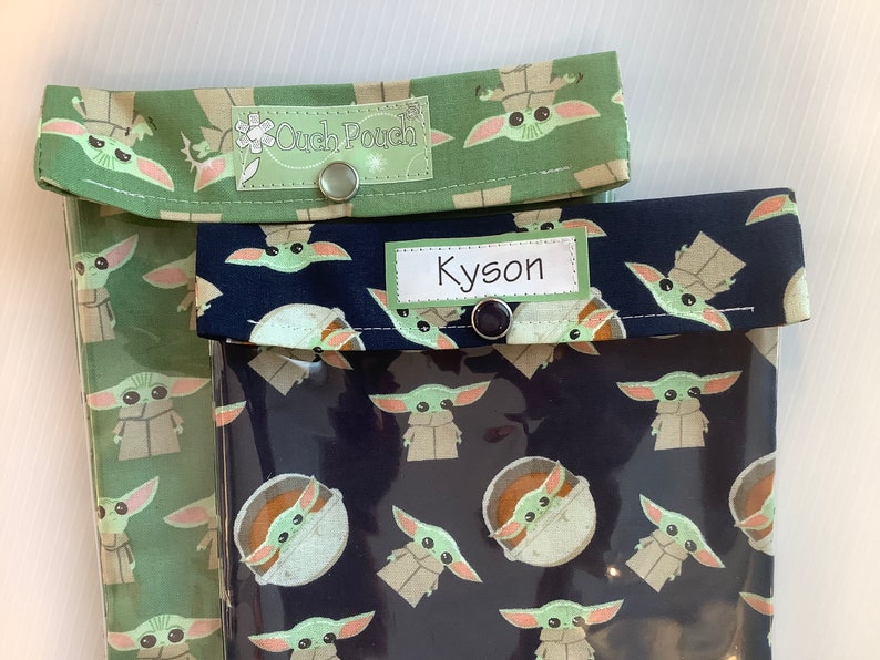 Puede incluir: Dos bolsas de aperitivos reutilizables con un frente de vinilo transparente y una parte posterior de tela. Las bolsas est&aacute;n decoradas con un patr&oacute;n repetido de un personaje alien&iacute;gena verde. Una bolsa tiene una parte posterior de tela verde con una etiqueta que dice "Ouch Pouch" y la otra bolsa tiene una parte posterior de tela negra con una etiqueta que dice "Kyson".