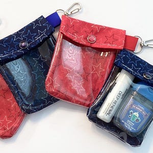 May include: Four small, clear pouches with star patterns in red and navy blue, each with a snap closure and a silver carabiner clip. One pouch contains a hand sanitizer and a lip balm.
