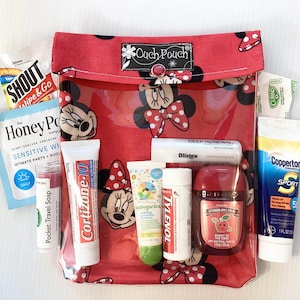Könnte beinhalten: Eine rote Reißverschlusstasche mit einem durchsichtigen Plastikfenster, die mit einem Minnie Mouse-Motiv verziert ist. Die Tasche enthält verschiedene Reisegrößen von Toilettenartikeln, darunter Sonnencreme, Lippenbalsam, Handdesinfektionsmittel und Lotion.