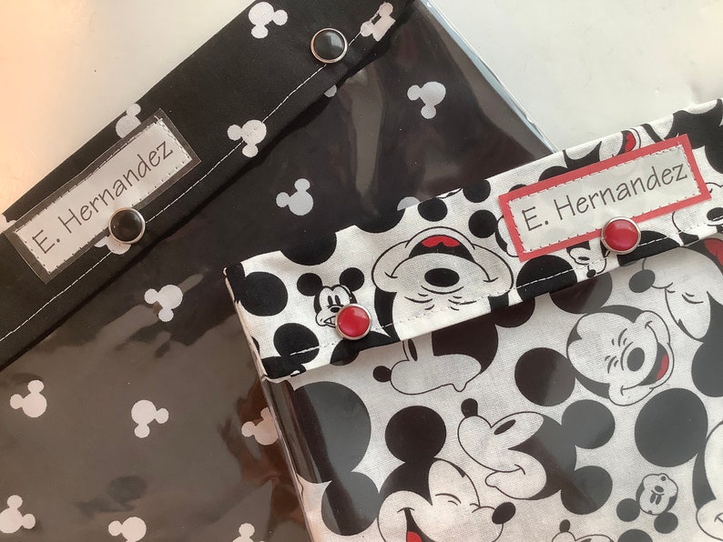 May include: Two black and white Mickey Mouse fabric pouches with clear vinyl front panels. Each pouch has a black label with white text that reads "E. Hernandez". The pouches have red buttons.