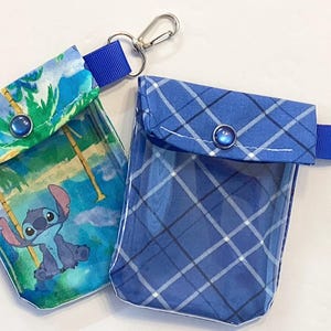 May include: Two small, clear pouches with blue accents and silver carabiner clips. One pouch features a Stitch character print against a tropical background. The other has a blue plaid pattern. Both have blue ribbon loops and snap closures.