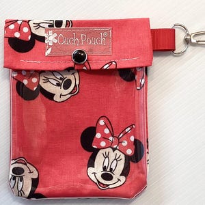 May include: Red fabric pouch with a clear plastic window, featuring a repeating pattern of Minnie Mouse. The pouch has a zipper closure and a small metal loop for attaching a keychain. The text "Ouch Pouch" is printed on the front of the pouch.